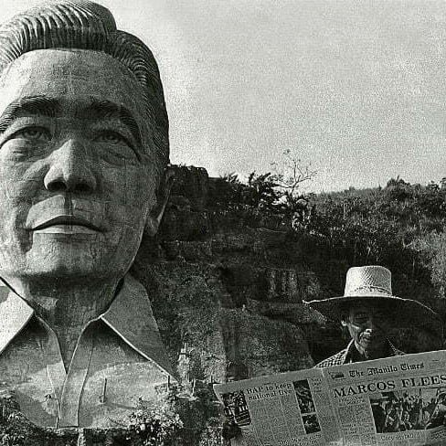 A farmer in Agoo, La Union, standing in front of the Marcos monument, reads about it the following day in a newspaper carrying the headlines “Marcos Flees.”